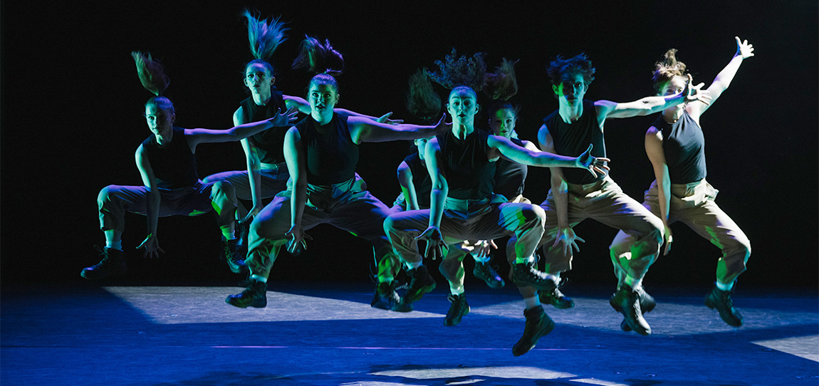 Eight college-age dancers perform on stage, leaping, with their hair flying above them. Their arms are out, fingers extended, and their knees are up. They are lit dramatically in green and purple hues.
